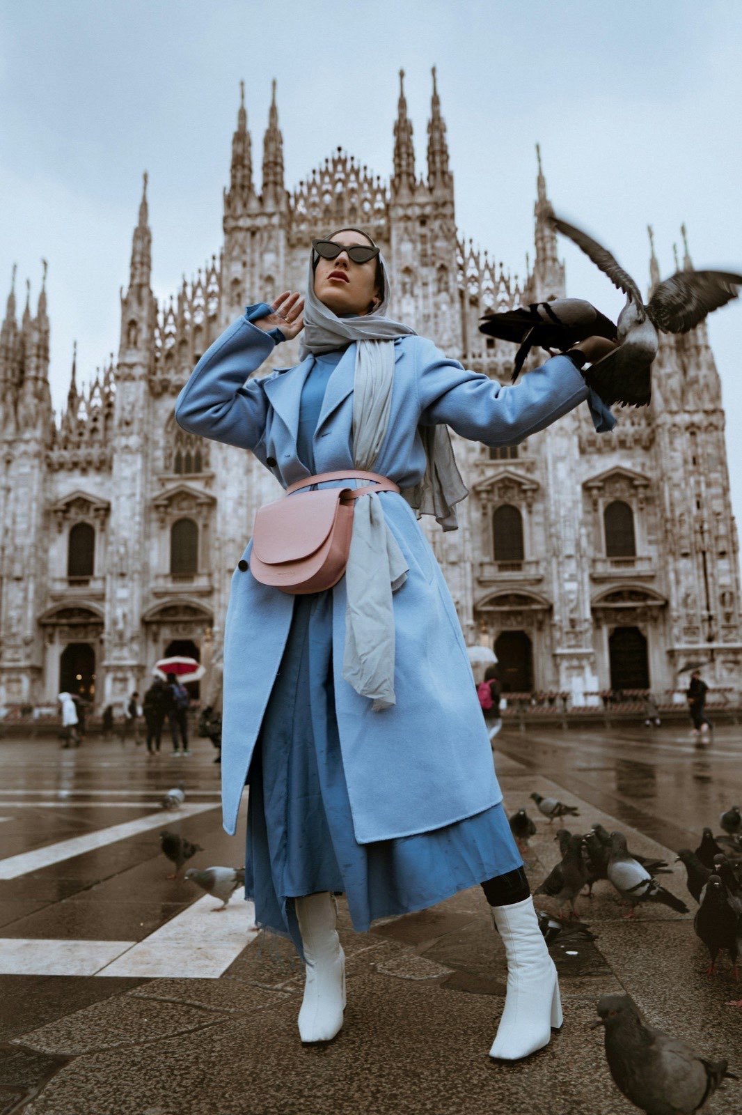 Woman in a light blue coat, scarf, and white boots standing in a city square
